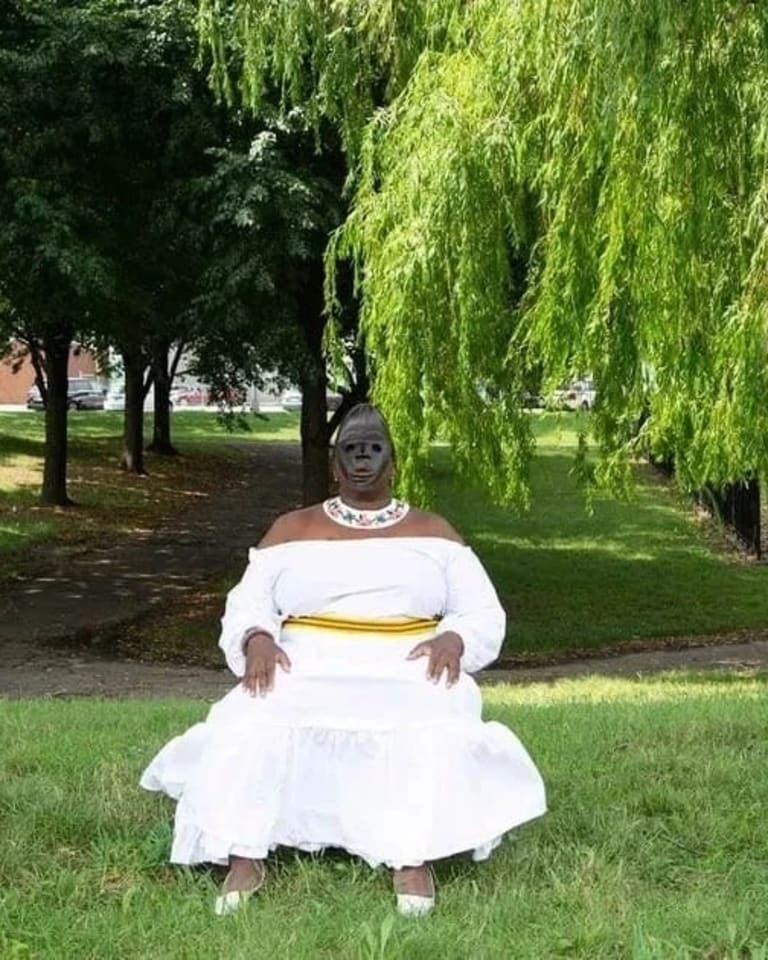 A woman in a white dress wearing a black mask sits in the middle of a green park space.