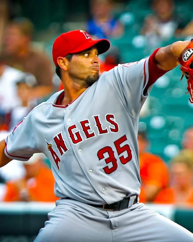 A man in a Los Angeles Angels jersey with he arm wound up to pitch a baseball. the crowd behind him is out of focus.