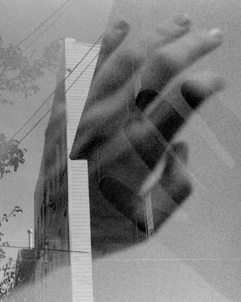 Black and white photo of two hands transposed over an apartment building with trees and power lines.