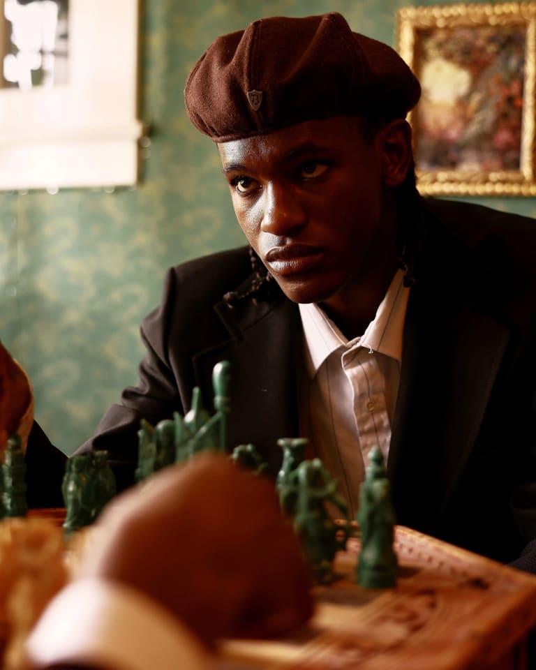 A dark skinned man wearing a beret sits on a velvet couch in a lavishly decorated room, he is bent over a game of chess and he appears to contemplate his next move while making eye contact with an unseen opponent