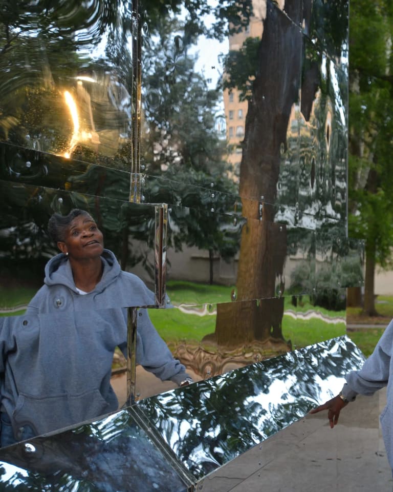 Image of a woman in a grey sweatshirt looking at her reflection in a mirrored sculpture in the park. The Battle Is Joined, Monument Lab, Mural Arts, Public art commission, Vernon Park, Philadelphia, PA, 2017