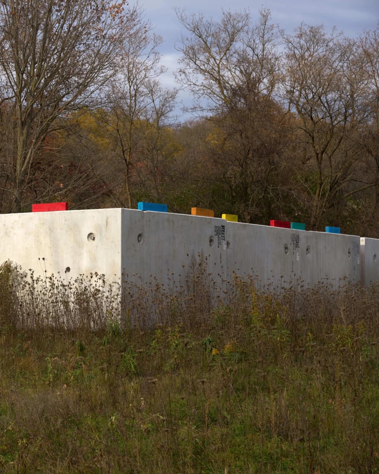 A photograph of a cement wall in a rural forest landscape. On top of the wall are multi-colored pieces of wood that make a repeating pattern.