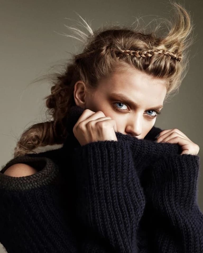 An image of a blonde woman with braided hair staring at the camera, covering the lower half of her face with her black sweater