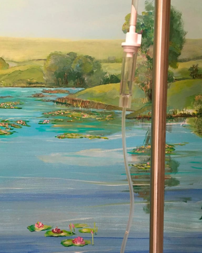 The background shows part of a wall on which is painted a mural of a landscape of a bucolic body of water with pink lotuses floating on it. The background of the mural is green and yellow hills and a bluish sky. In front of the mural, dissecting right of center, is part of a copper pole, from which hangs part of a plastic tube with liquid dripping through it.