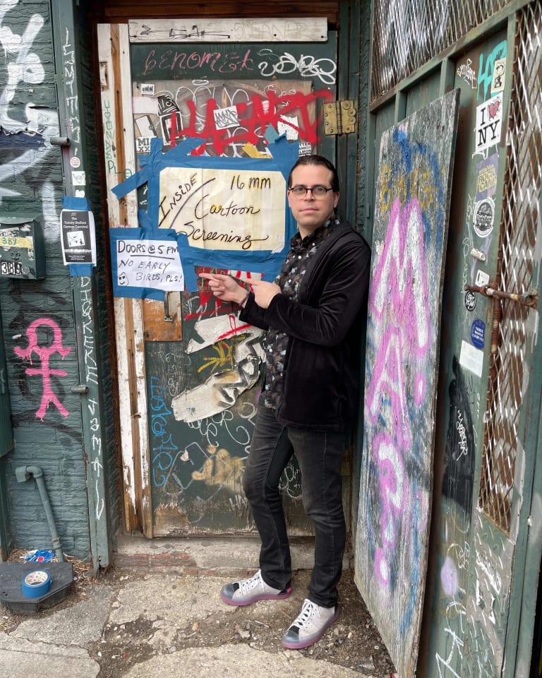 Picture of a man standing in front of a door covered in posters and graffiti, pointing to a poster that reads "Inside: 16mm Cartoon screening. Doors at 5pm, no early birds."