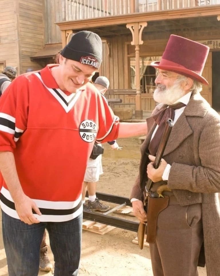 Two men on a movie set of a western town. One of them is in casual, modern clothes and the other one is dressed up to fit the set of an old western town, with a red top hat and a shotgun in his hand.