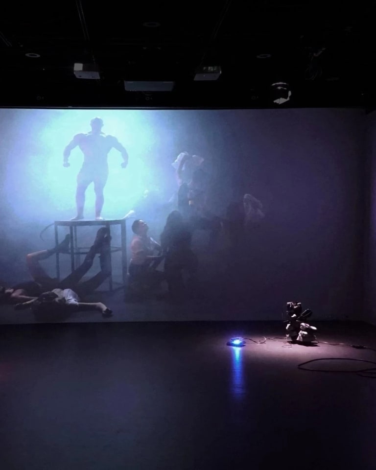 Picture of an installation with three walls being projected with the same image: A foggy dimly lit environment, with several people strewn about the floor, and a single figure of a strong man standing, backlit, against the only source of light.