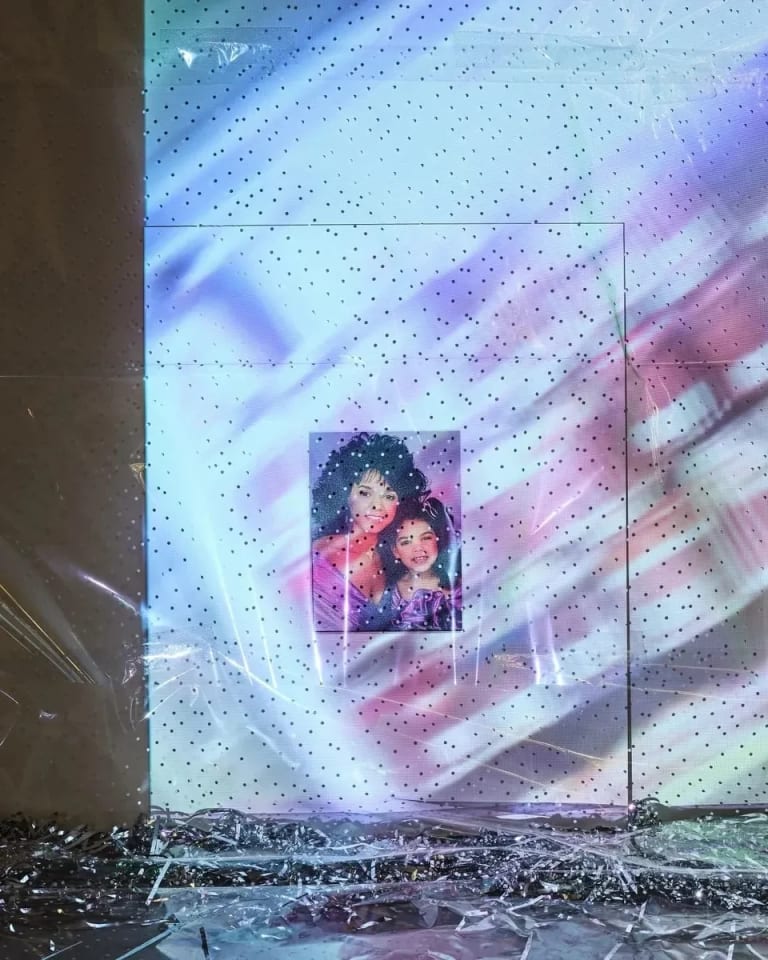 a painting of a woman and young girl in a white installation space, shrouded by cellophane and projected colorful images 