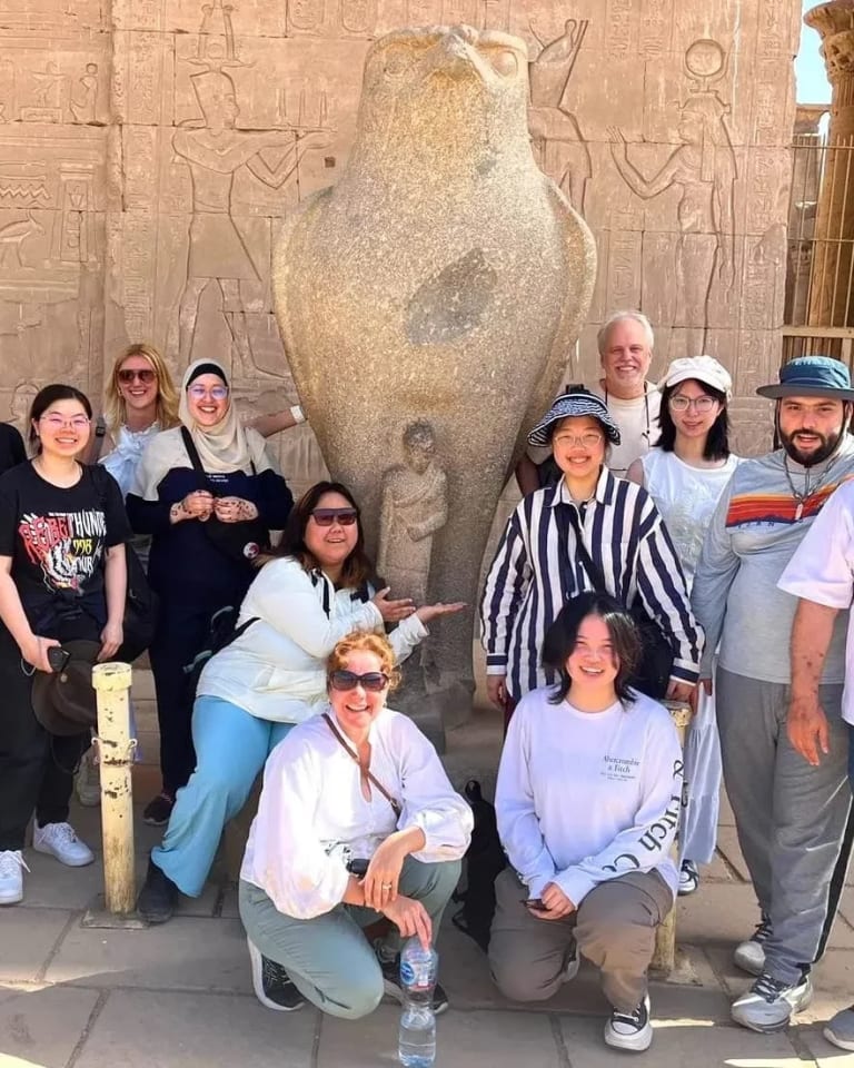A group of students and faculty members, some wearing sunglasses and hats stand and pose in front of a ancient building in Egypt. 