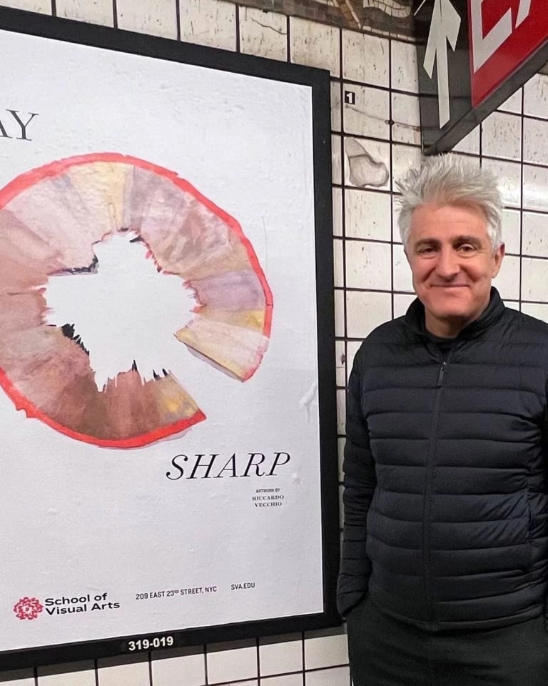 A man with gray hair, wearing a black jacket, smiling as he stands next to a poster in the subway.
