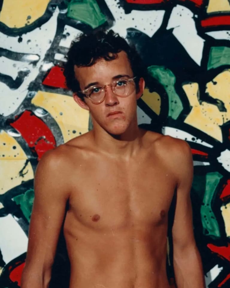 A young Keith Haring, shirtless and in front of a colorful mural. 