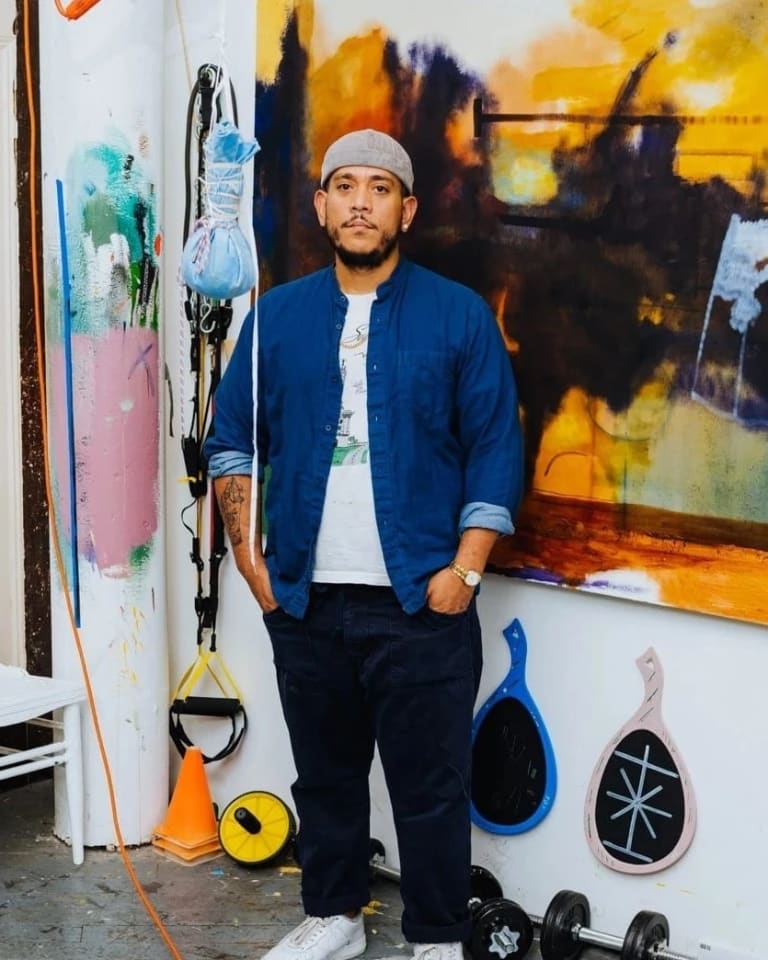 A photograph of Kenny Rivero in his artist studio. Kenny is standing in front of a wall with a paintings and other objects hanging on the wall. Kenny wears a white shirt with a blue button-down over it and black pants. His hands are in his pocket and he is looking at the camera.