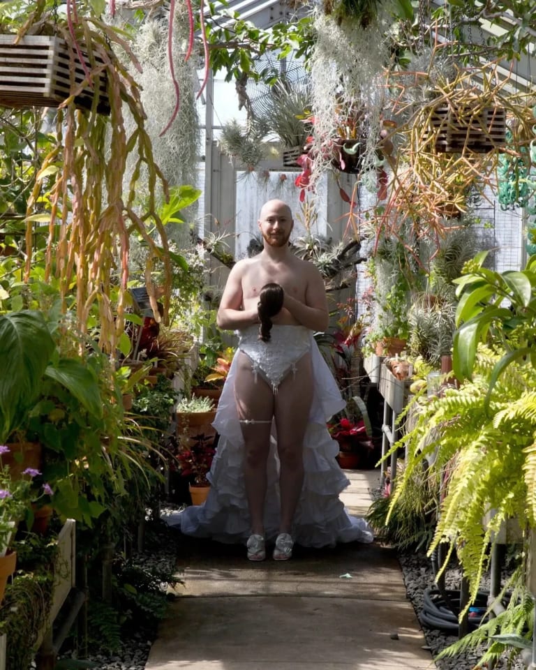 A transman standing in the middle of a greenhouse wearing high waisted, embellished white underwear with a bustle/train attached, and a garter, holding a wig in their hands like a bouquet. It looks like they are about to walk down the aisle.