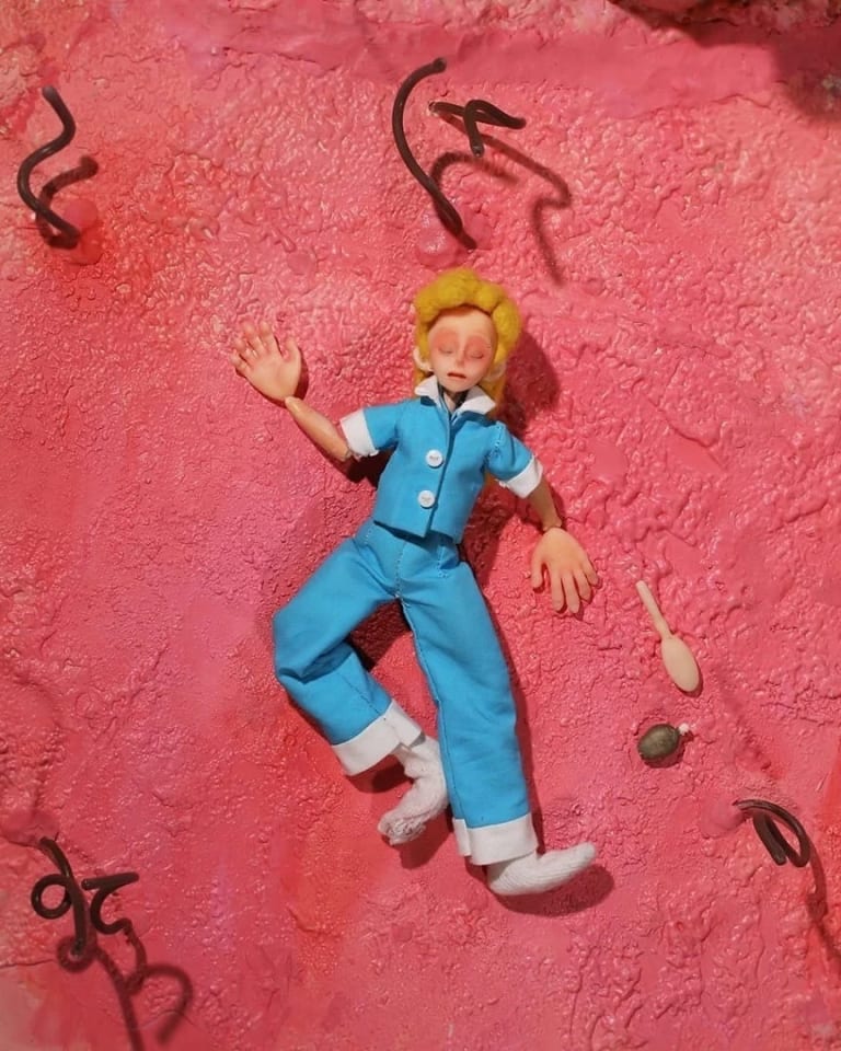 Still from a stop motion film depicting a blonde female character in blue clothes laying unconscious in a fleshy pink floor with neon green goopy substances around her and swirly hair-like things coming from the floor. 