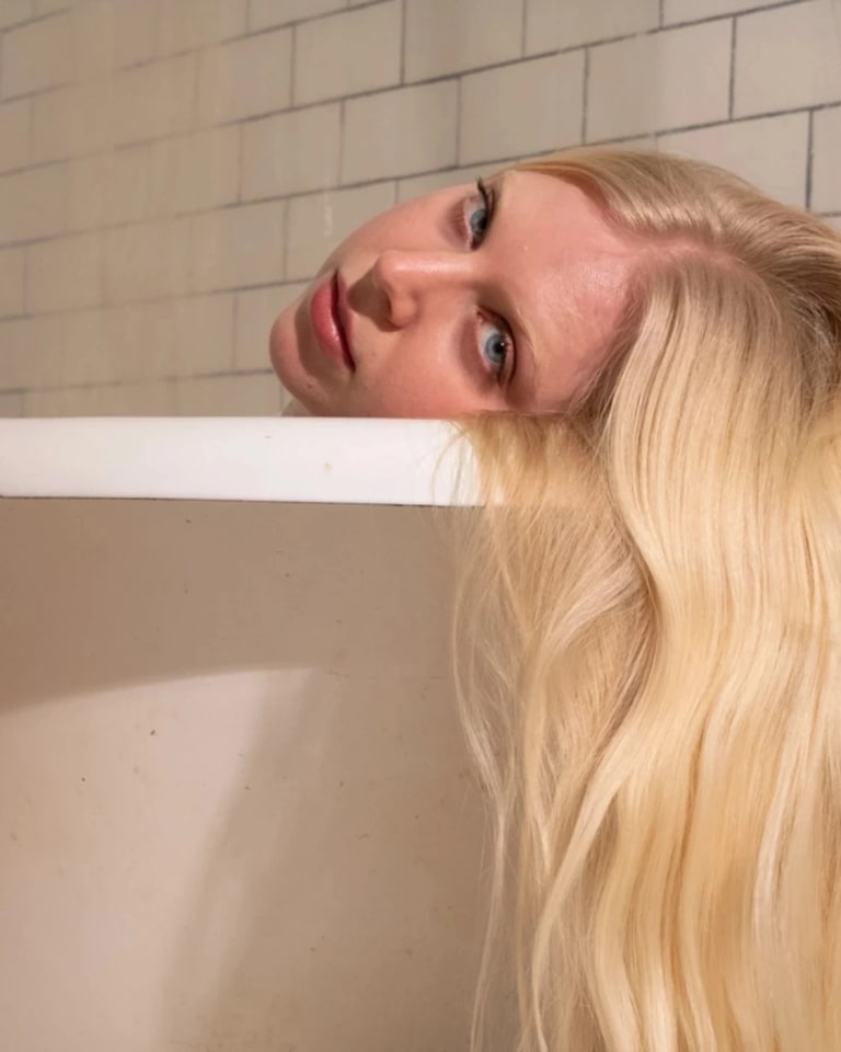 screenshot showing the title “Maggie Dunlap Wants You to Imagine the Worst” in black block letters on white at to the right, and on the left a white bathtub viewed at eye level, with a blonde woman resting her head on the tub edge and looking at us. Her straight hair hangs down over the tub’s edge at the right of the image.