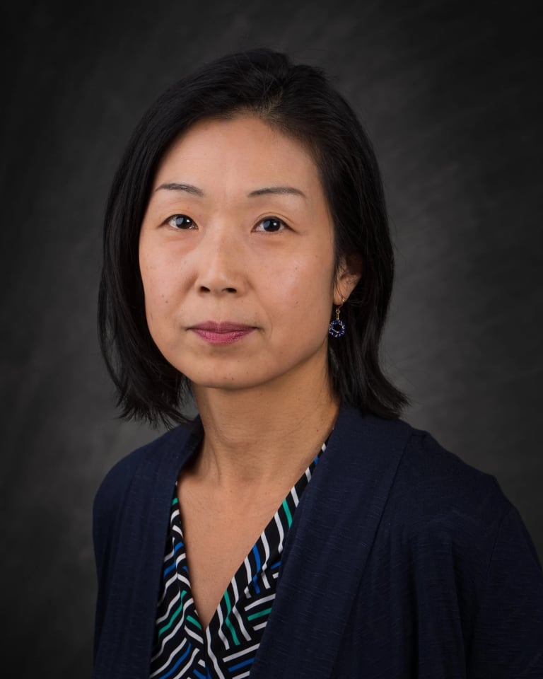 A headshot of Midori Yoshimoto. Midori is wearing a stripped shirt with a navy sweater. She is set against a gray background and is looking at the camera..