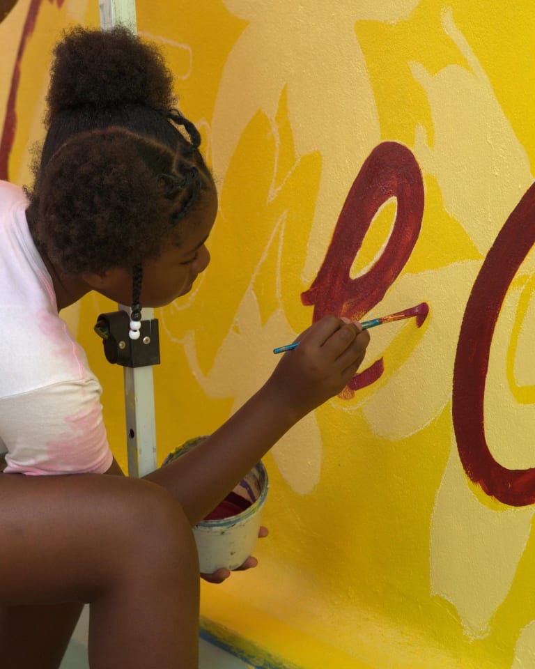A person in a t-shirt in shorts sits on a chair, crouching to paint a yellow wall with red paint