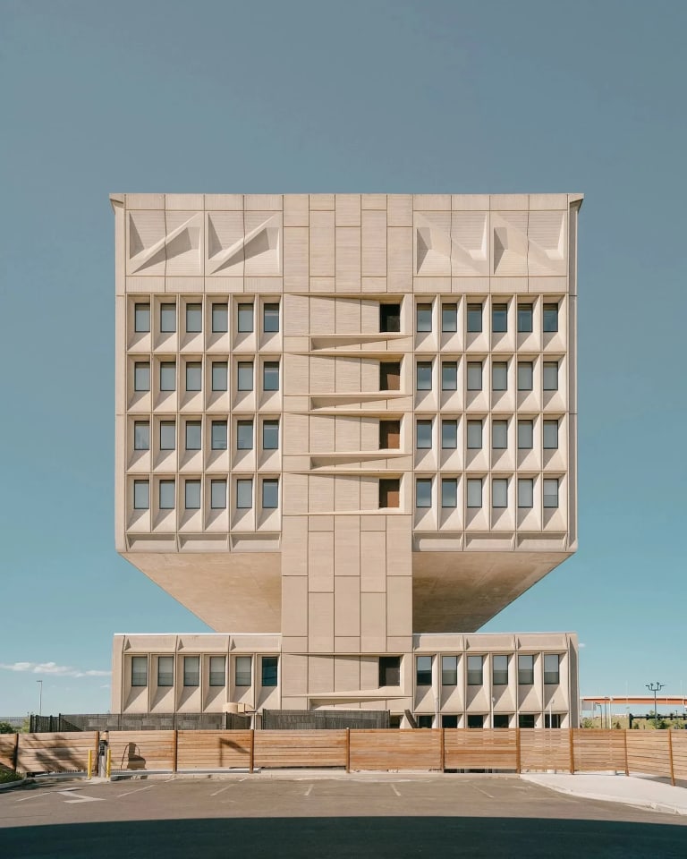 architectural color photo of a large square concrete building against a blue sky.