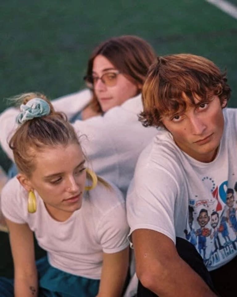 Three teens wearing white T-shirts sit on a football field in a cluster with moody looks