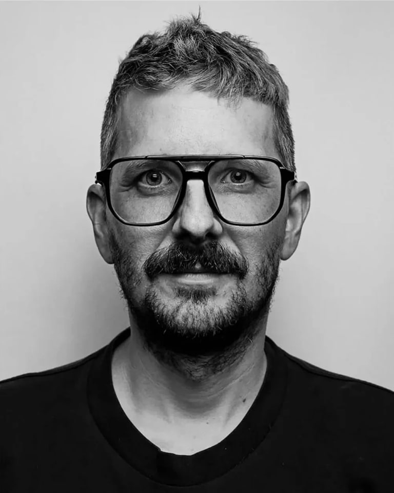 A black and white photo of a man in a black shirt with grey hair wearing black glasses staring straight ahead at the camera.