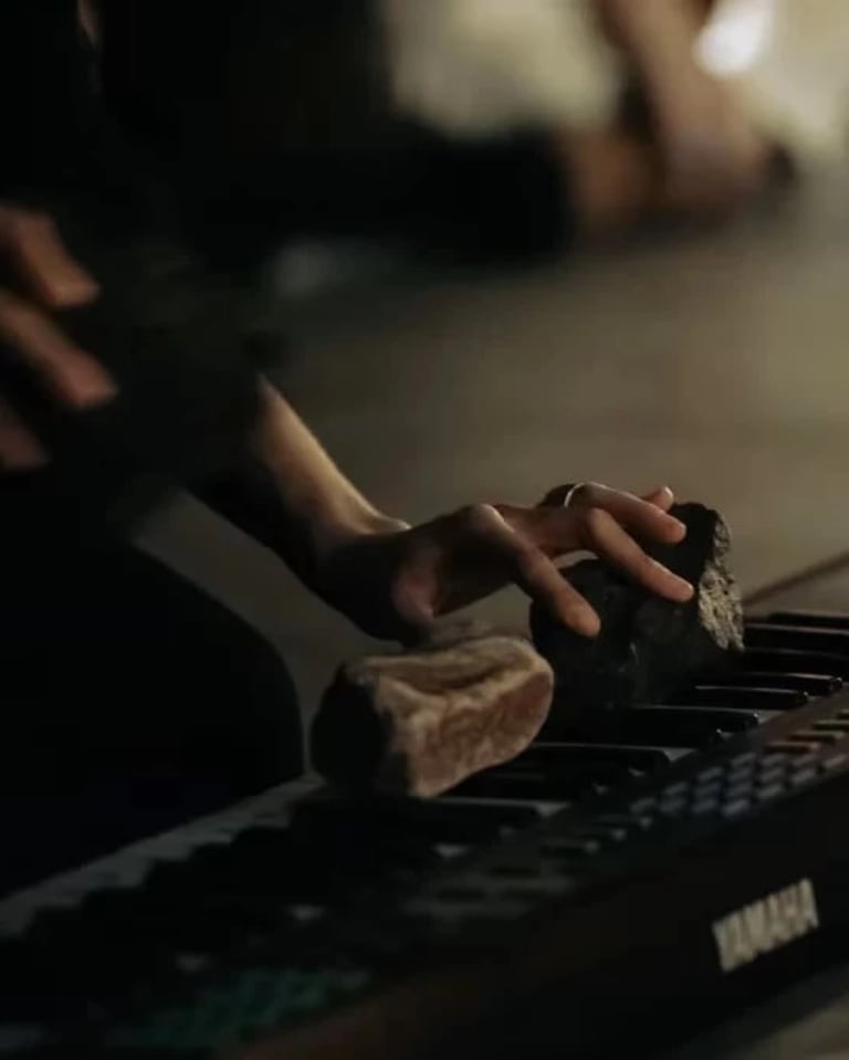 Photographic image of a hand on a keyboard. The keyboard has two rocks on it that the hand is pressing into the keys