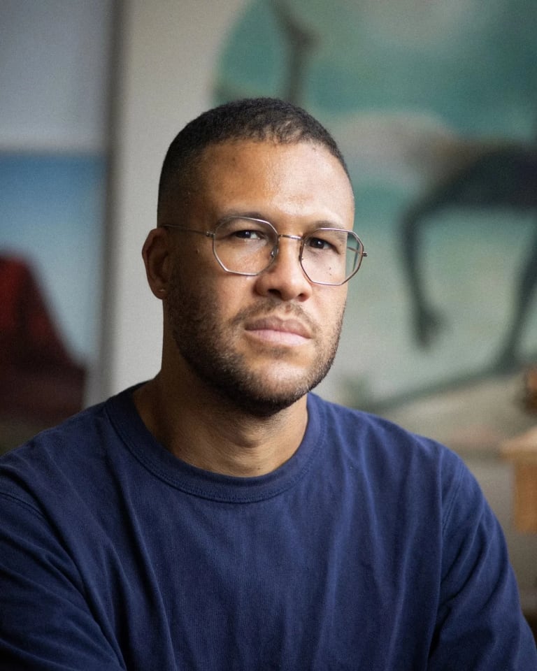 A photograph of Simon Benjamin. Simon is wearing a navy blue shirt with thin wired glasses. He is looking directly at the camera with a relaxed expression. Some of his artworks are in blurred in the background.