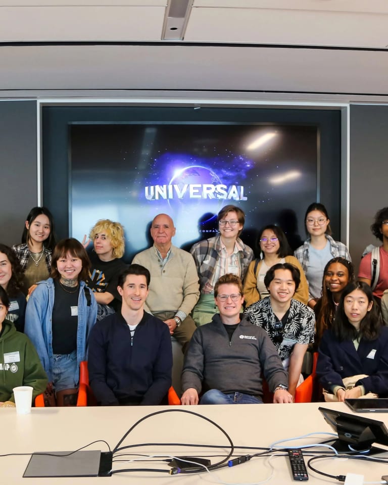 A group of students and adults, likely artists, organized in three rows and posing in front of a screen that displays Universal Studio's name and logo.