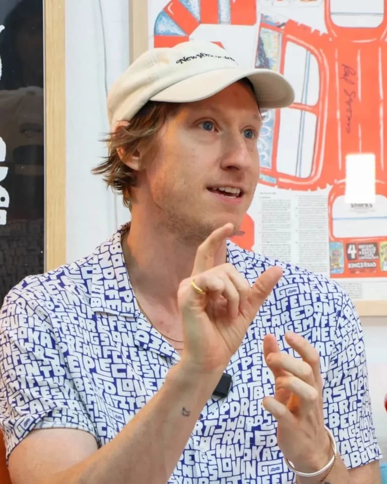 A man in a black and white shirt wearing a white baseball cap sits on a couch holding his figures apart with abstract paintings behind him on the wall.