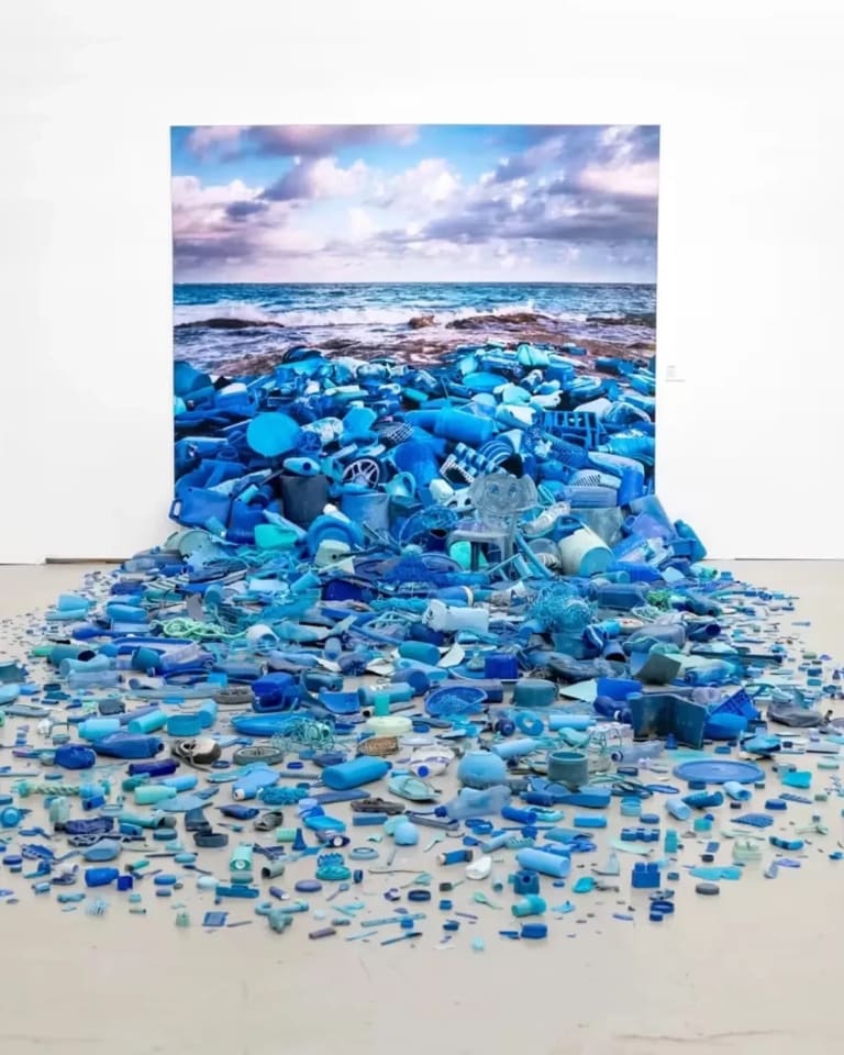 A gallery space with a landscape picture on a wall and blue pieces of plastic spilling out onto the floor.