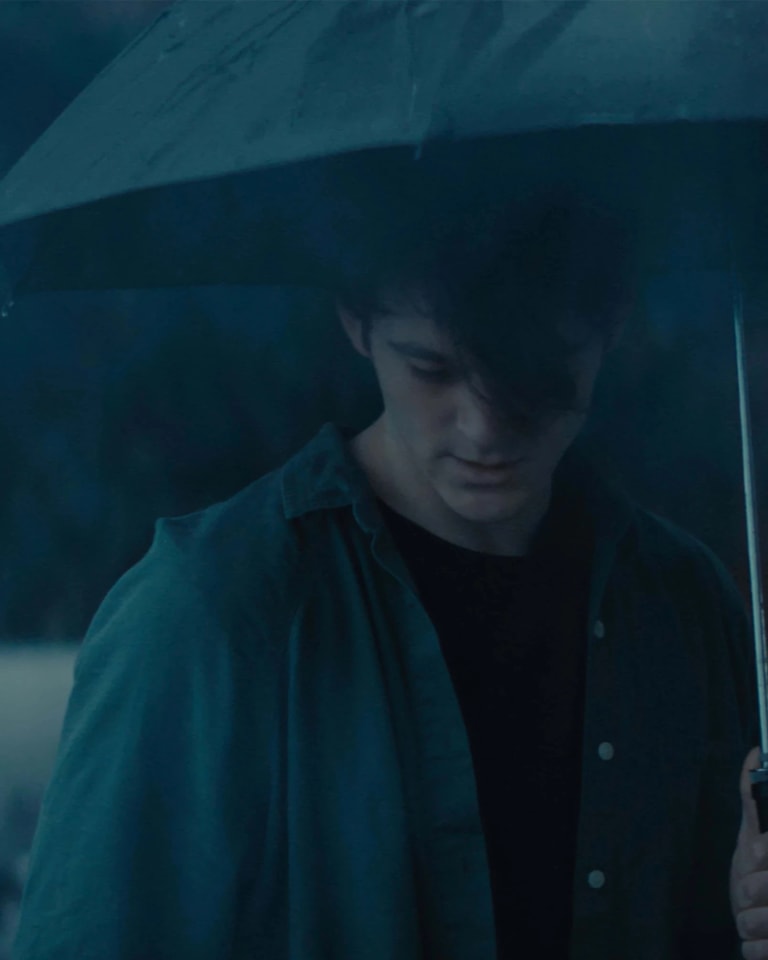 Film still of a man under an umbrella, looking down at the ground. The colors are all very muted and cool.