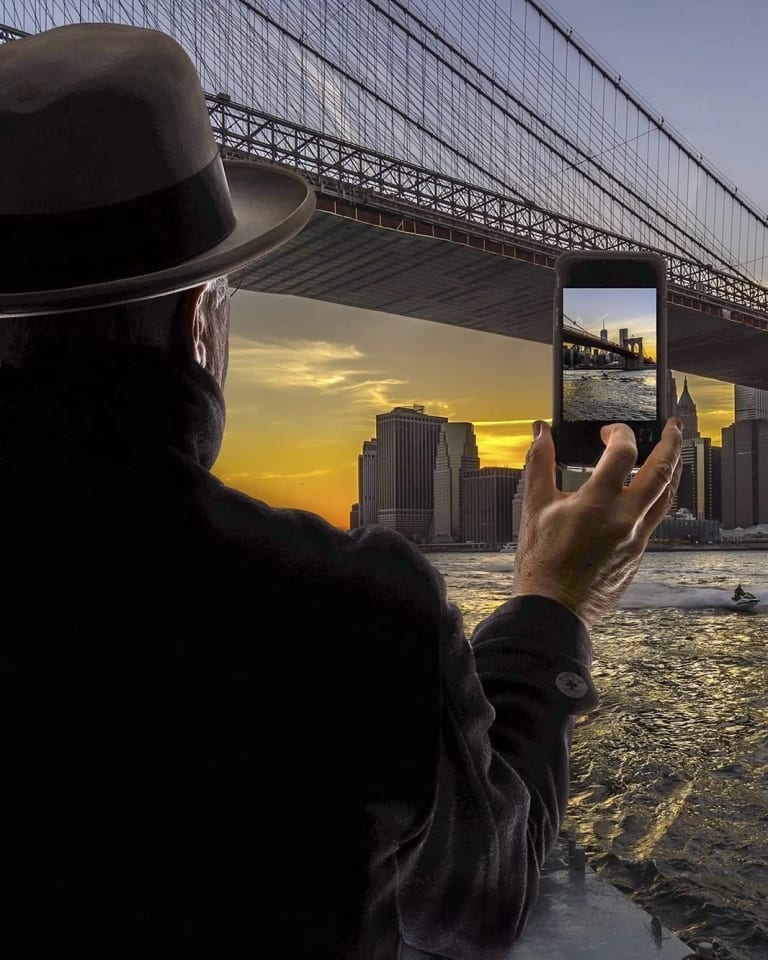 A man, wearing a brown fedora and a black coat, whose back is turned toward us, is looking across the Brooklyn Bridge at the Lower Manhattan skyline against the sunset. His right hand is raised as he is taking a photo of the skyline on his cell phone.