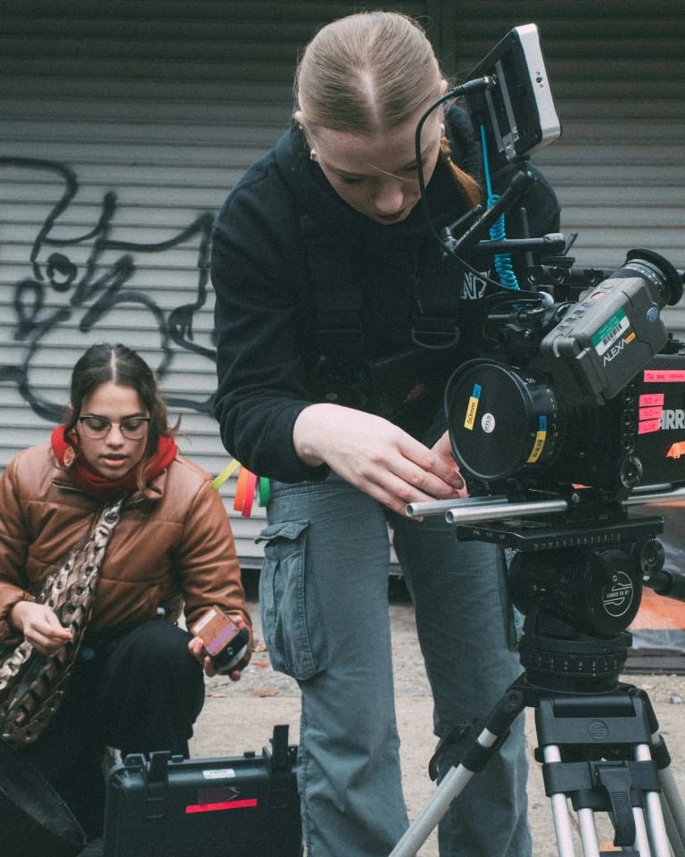 The individuals stand in an outdoor urban setting. The one on the far right is a blonde woman who is wearing a black hoodie and adjusting a large film camera. The other two are working a sound machine and wearing outdoor coats. 