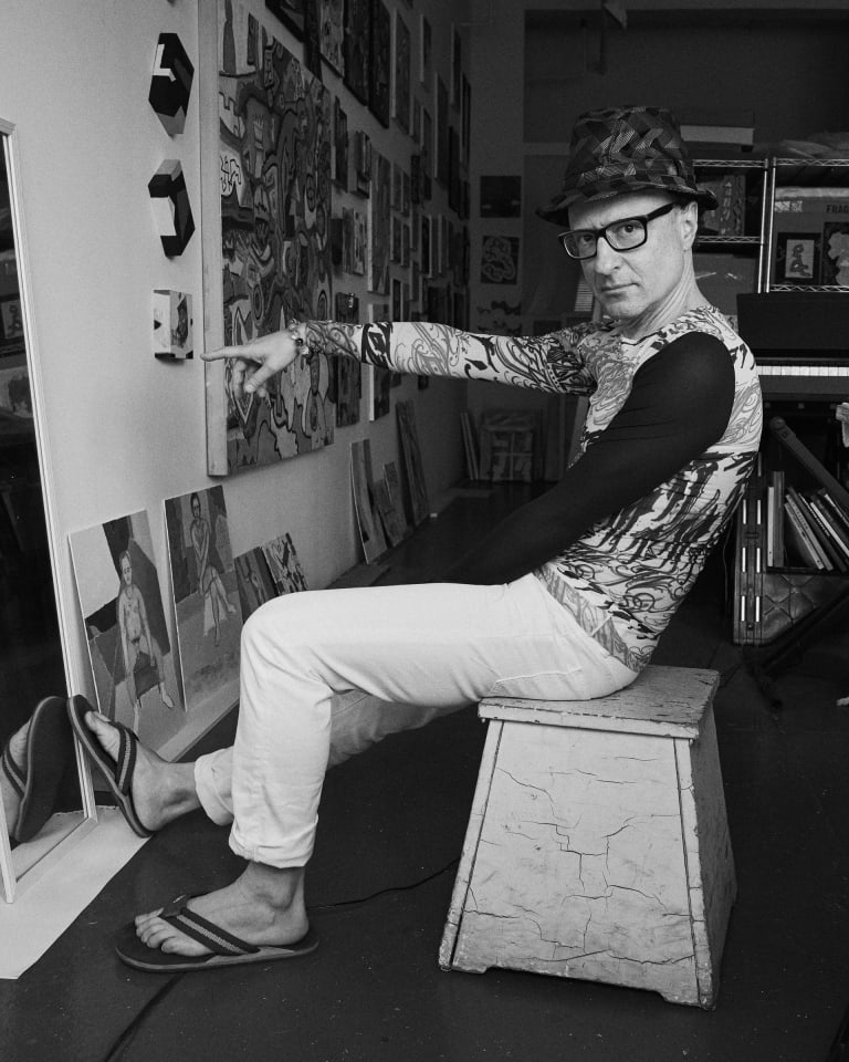 Black and white photo portrait of the artist Wayne Koestenbaum pointing at a painting in his studio.