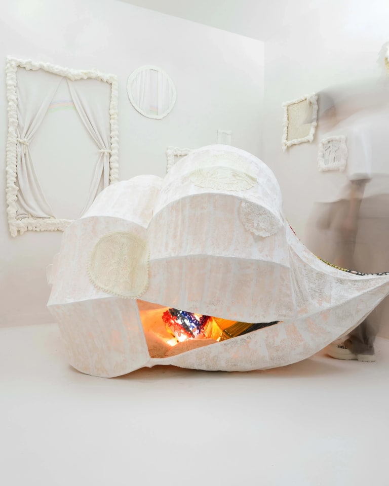 An installation in a small white room of a white cloth pod, warm light glowing within, and empty frames of white fabric on the walls.