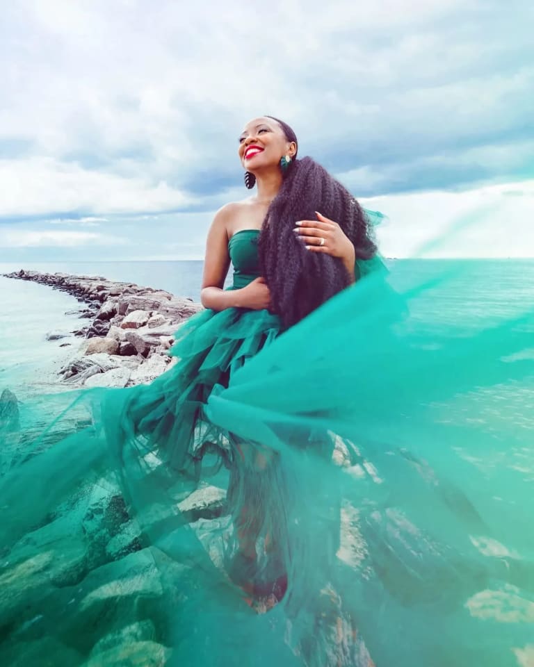 A woman in a teal dress stands smiling on a rock jetty, surrounded by the water.