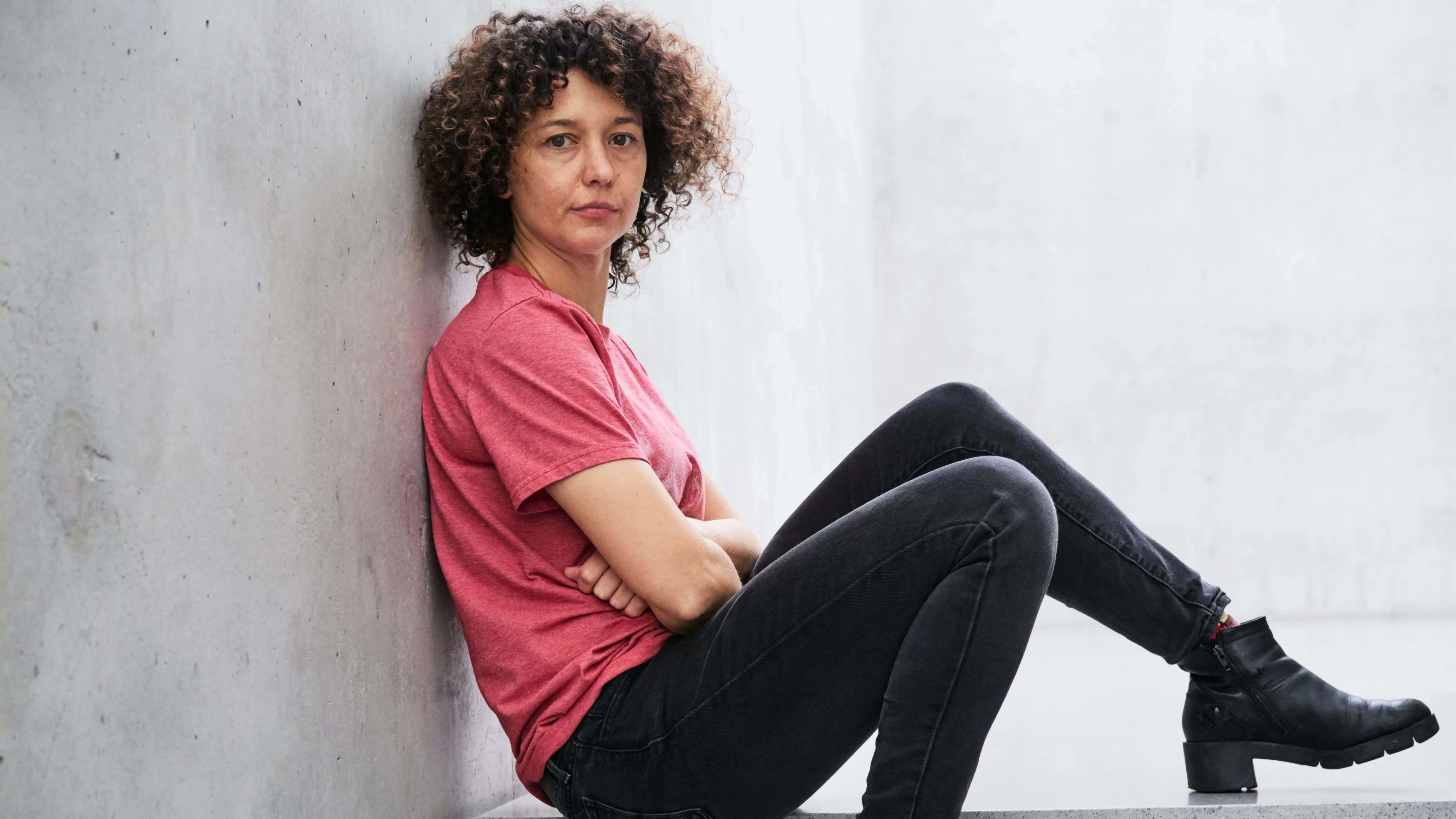 A photograph of Mika Rottenberg. Mika is sitting on a cement step with her back against the wall. She is wearing a red t-shirt with black jeans and boots. Her head is turned to the camera with a relaxed expression.