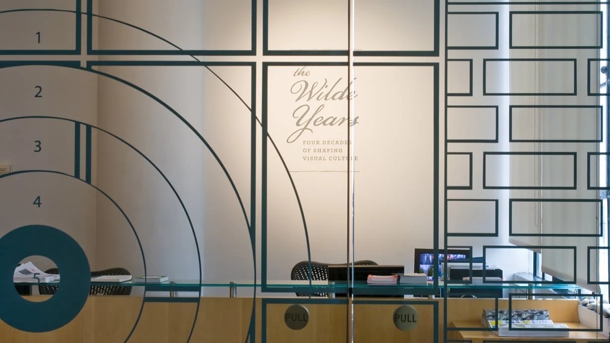 Photograph from outside SVA Chelsea Gallery glass doors, looking inside, with exhibition title visible behind desk.