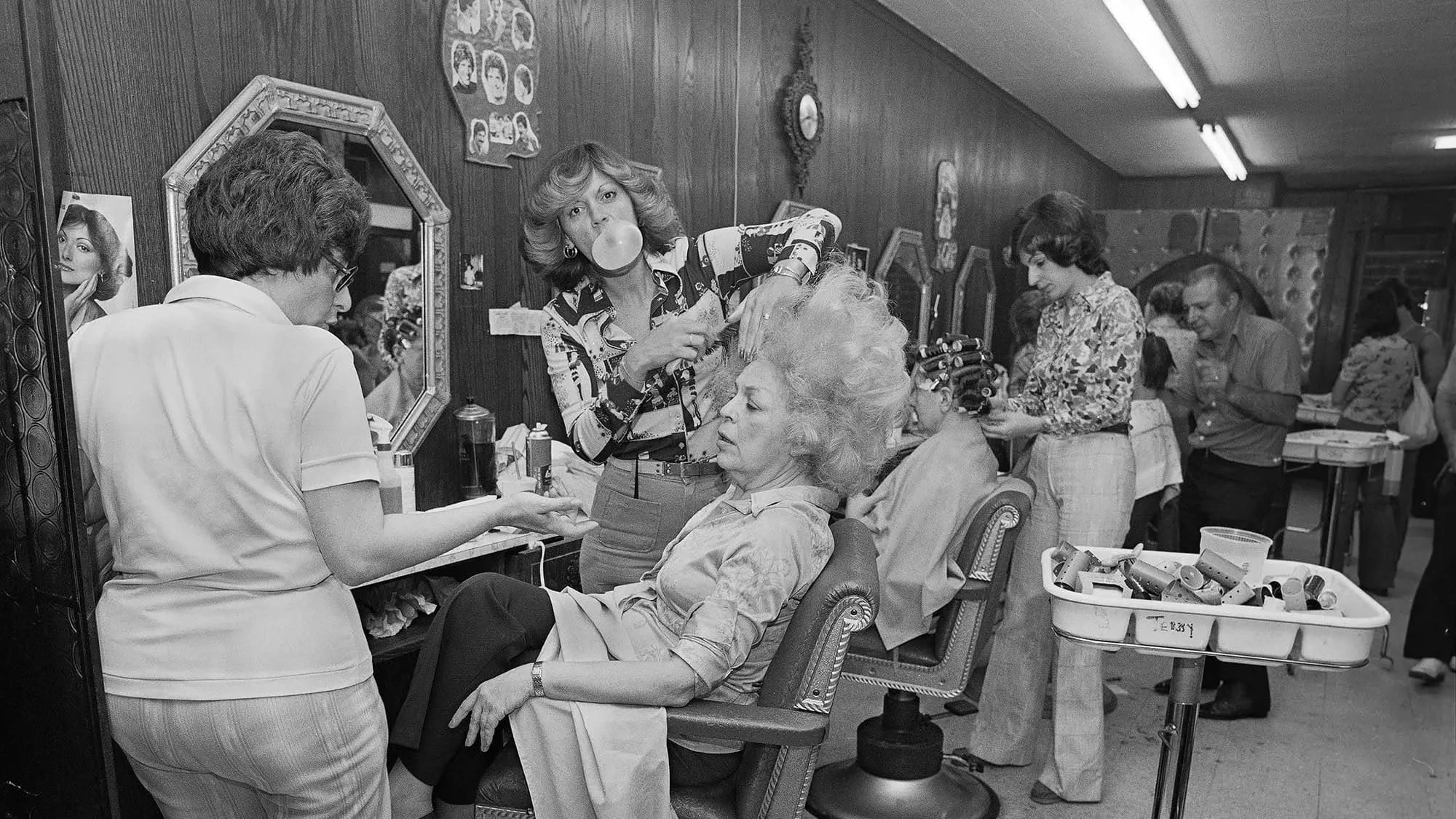 Artist’s mother in a beauty salon getting her hair teased while the beautician blows bubble gum.