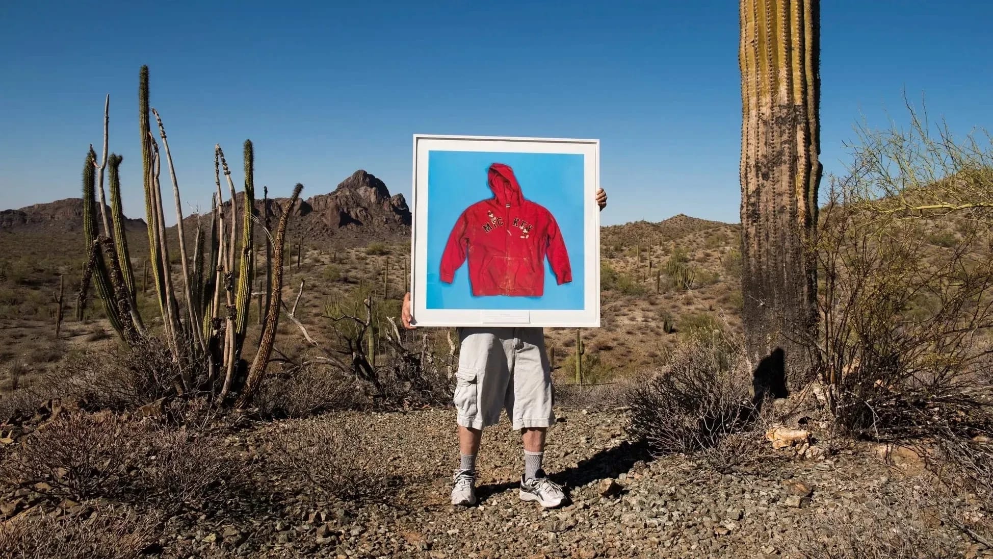 Photographer Tom Kiefer near his studio in in Ajo, Arizona, in the desert with a huge cactus next to him. He is holding up one of his framed photos of a red hooded sweater on a blue background. The letters on the sweater say MICKEY.