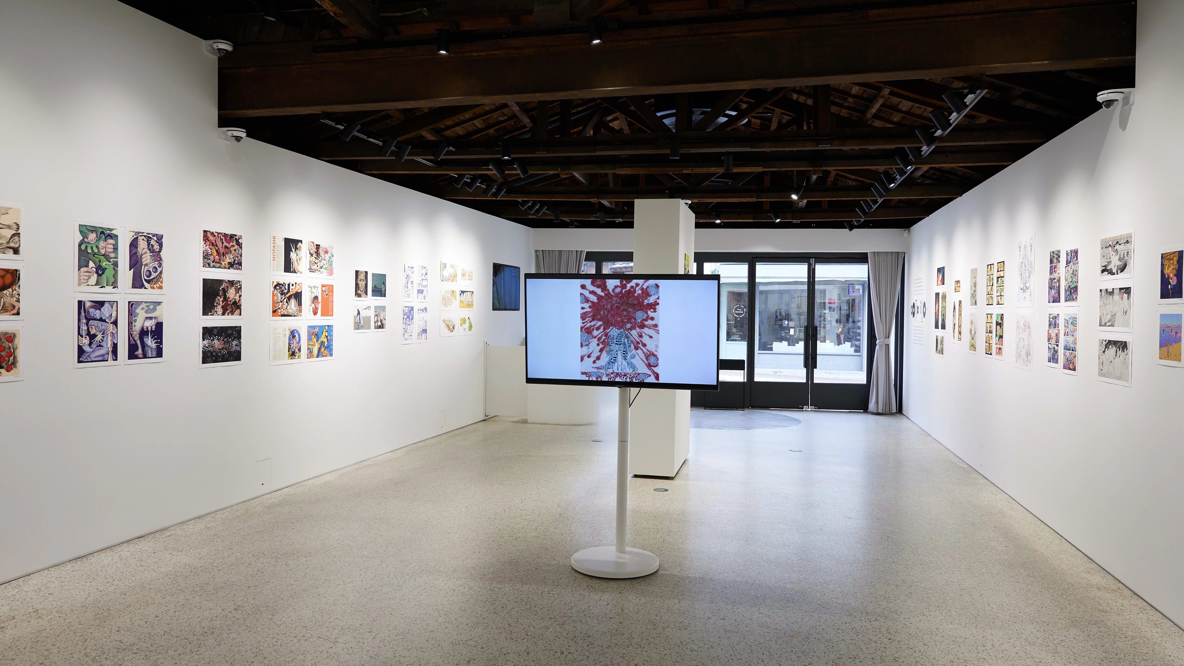 Gallery installation displaying colorful comic and illustration-style artworks arranged linearly along both walls. A large monitor on a white pedestal at the center shows a figure with red abstract patterns. The space features an open ceiling with exposed dark wooden beams, track lighting, and natural light filtering through glass doors at the far end.