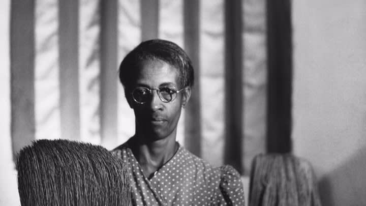 A Black woman stands in front of the American flag holding a broom and a mop.