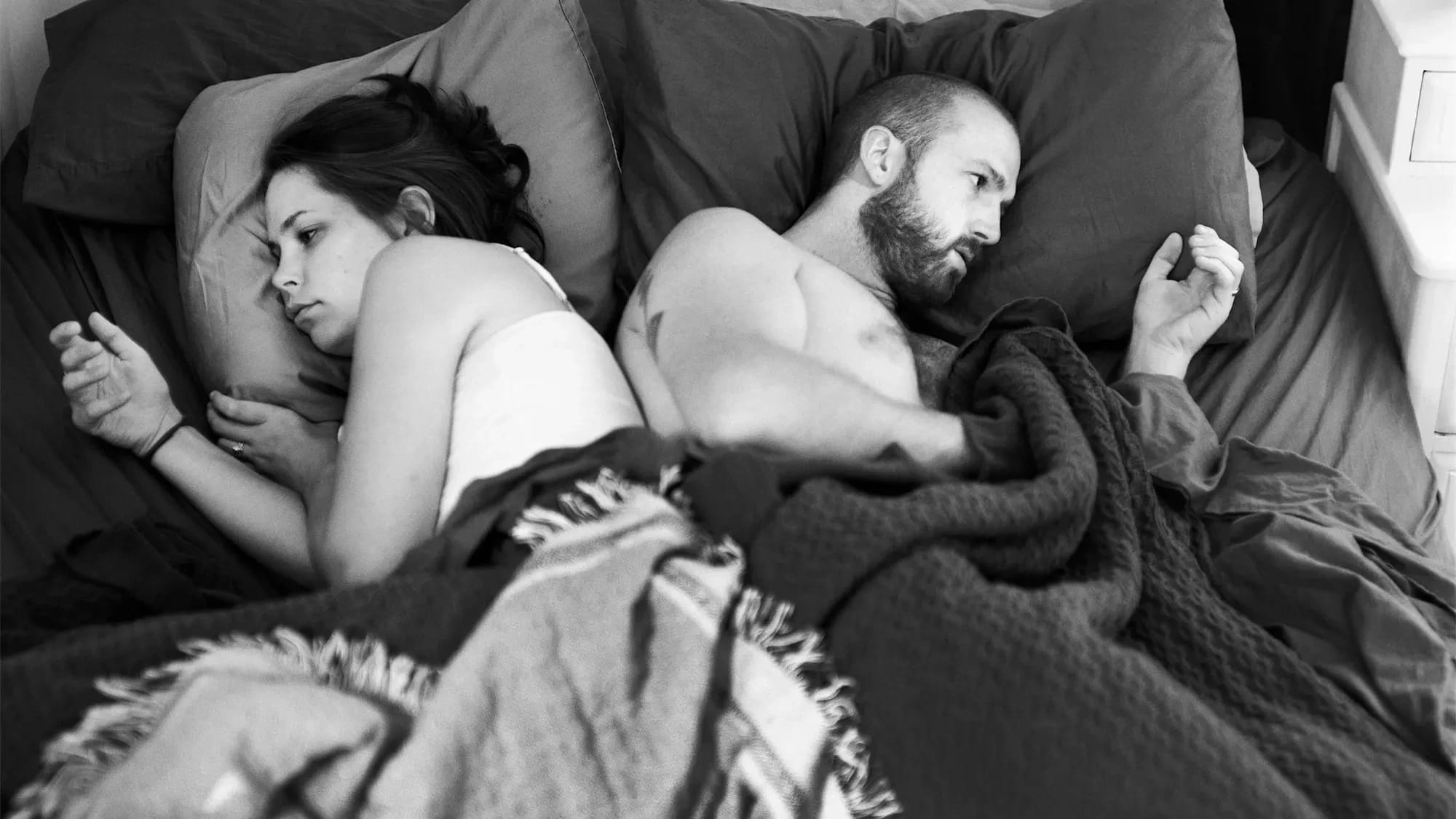 A black and white photograph of a man and a woman lying side by side in bed, facing away from each other. Each holds a hand in the air as if scrolling or holding a phone, though no devices are present. Their expressions are distant and disconnected. The photo is black and white and emotionally charged.