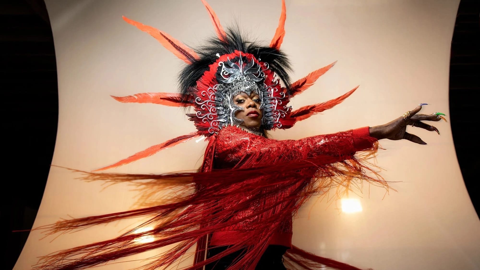 A studio portrait of a person in an elaborate headdress and red shirt.