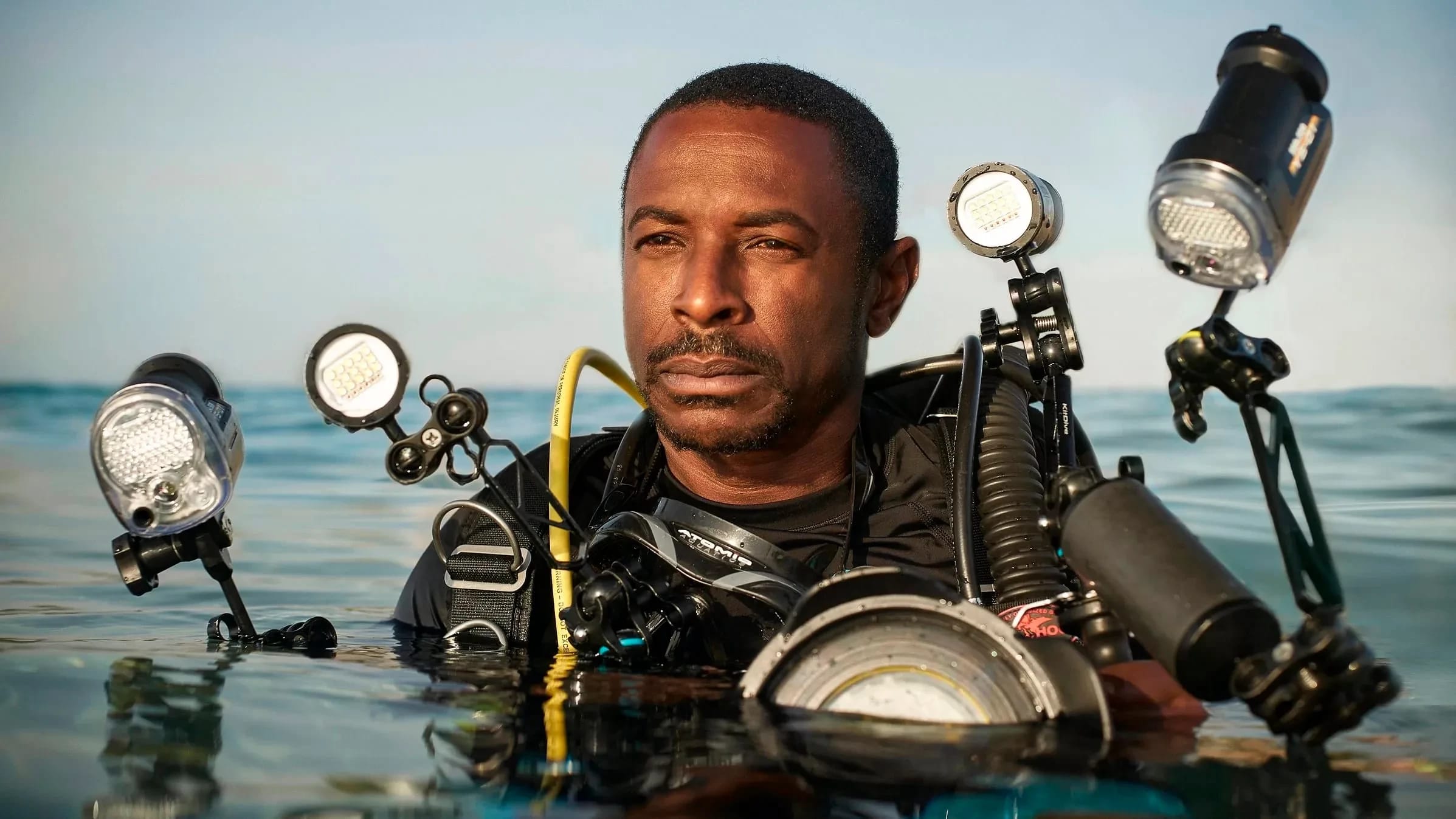 A portrait of a scuba diver in water. He has darker skin and short hair, is wearing a black top, and is equipped with numerous lighting and camera equipments strapped to his body. His body is submerged in water from the chest down.