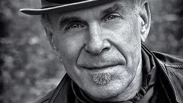 A black and white photo portrait of the photographer Douglas Dubler. He has light skin, is wearing a fedora & a leather jacket over a black top with a silk scarf around his neck.