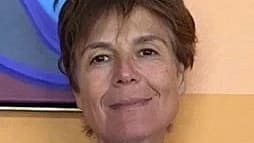 a person with short brown hair and brown eyes smiles in front of a beige wall 