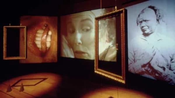 An image of a dark room looking at a diagonal angled wall with several spot lit frames hanging. The frames contain close up portraits. 
