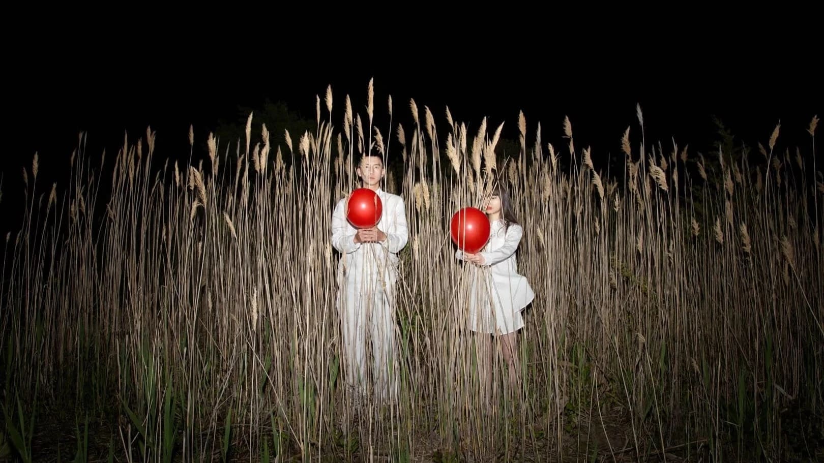 A man and woman wearing all white are each holding a red balloon, standing in tall reeds at night.