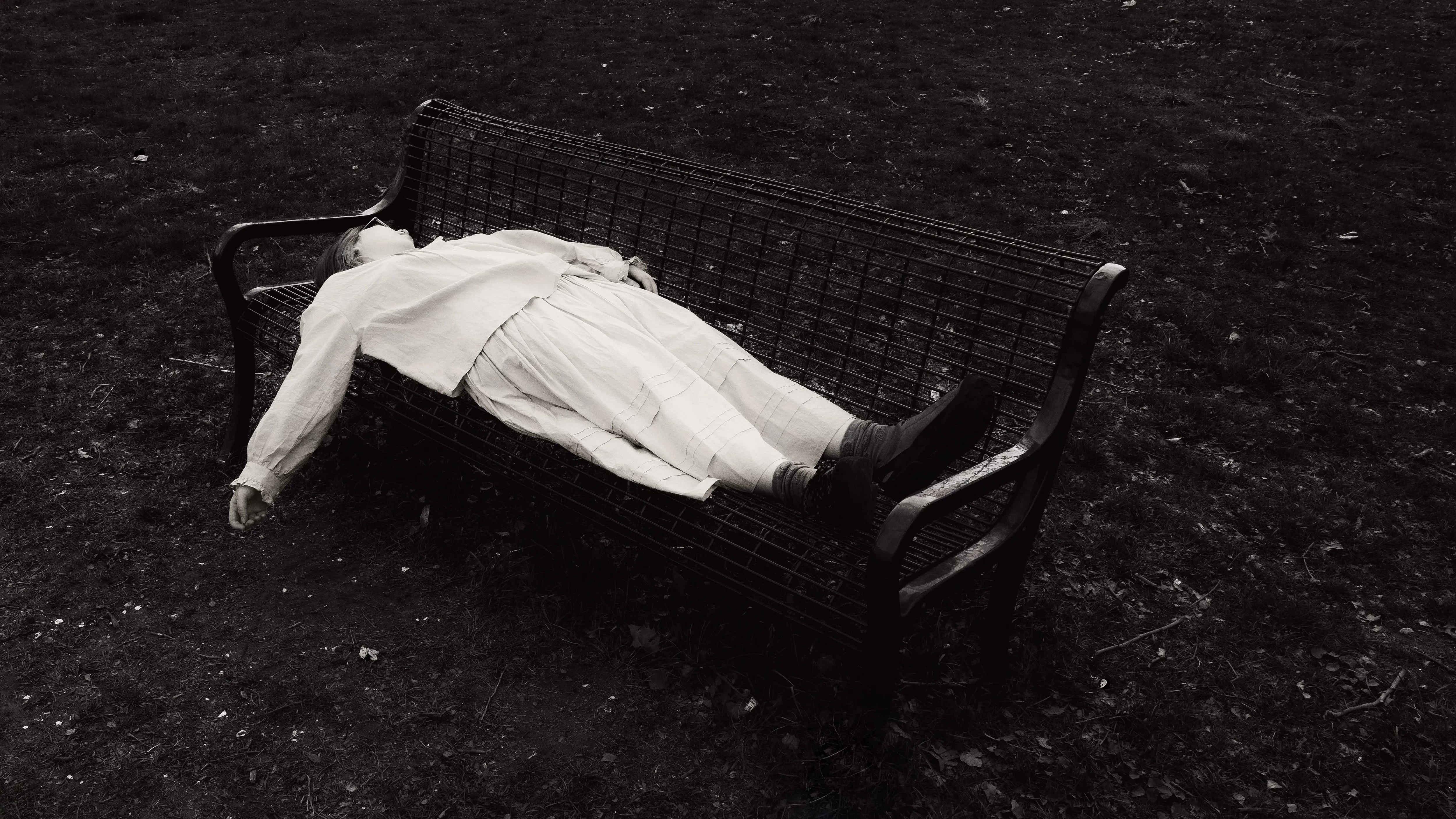 A black and white photograph of a girl lying on a bench, her hand hanging loosely, face obscured. The bench and her body are surrounded by chaotic bundles of grass.