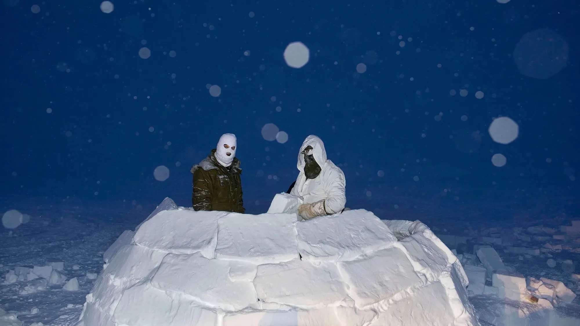 A color photograph of Two soldiers, wearing masks in an igloo made of snow blocks, at dusk while it snows, lit with flash.